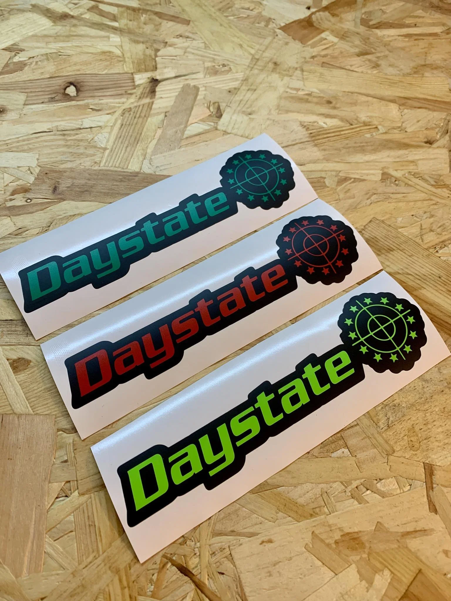 Three stickers with the word "Daystate" in different colors: blue, red, and green, each with a black target symbol inside a black circular design, placed on a wooden surface.