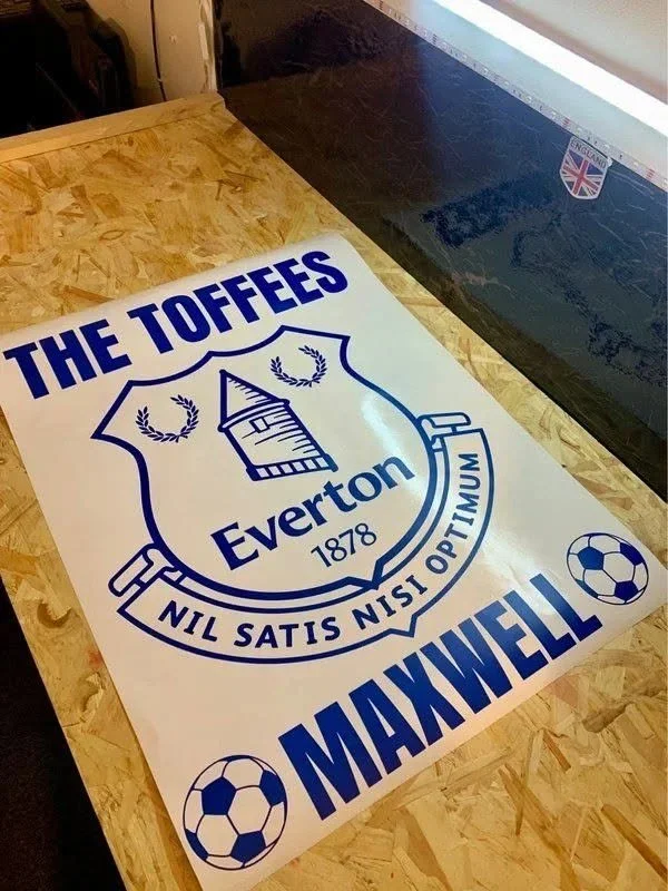A promotional poster for Everton Football Club, also known as The Toffees, with the motto 'Nil Satis Nisi Optimum' and the name Maxwell, lying on a wooden table.