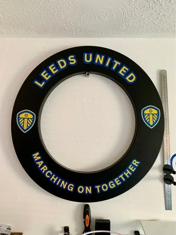 A large black circular sign on a white wall with yellow and blue text reading 'LEEDS UNITED' at the top and 'MARCHING ON TOGETHER' at the bottom. The sign features two Leeds United logos on either side, which are shield-shaped with a yellow background and blue details. There is a ruler with an orange and black saw on the right side of the sign, and some other objects are visible below.