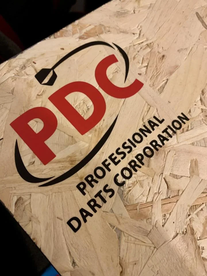 A die-cut sticker with the PDC logo and the text 'Professional Darts Corporation' on a wooden surface.