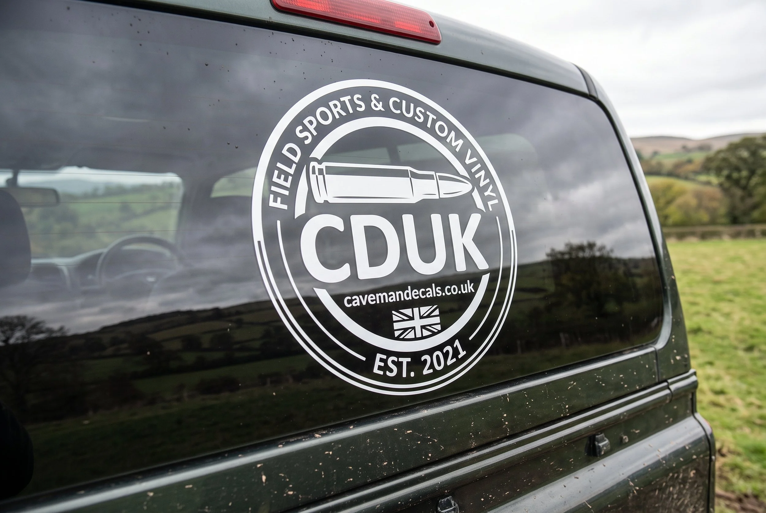 Black vehicle with a circular decal on the rear side window. The decal reads 'FIELD SPORTS & CUSTOM VINYL' and features a bullet illustration. The decal also includes a website address 'cavemadecals.co.uk' and an establishment year 'EST. 2021'. The vehicle is outdoors with a background of open fields and cloudy sky, and some dirt or water droplets are visible on the window.