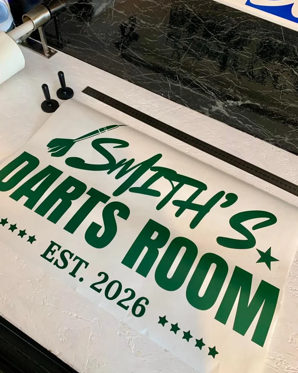 A printed sign with green text that reads "Smeltzer's Daughters Room," a feather graphic, and, "EST. 2026," on a worktable.