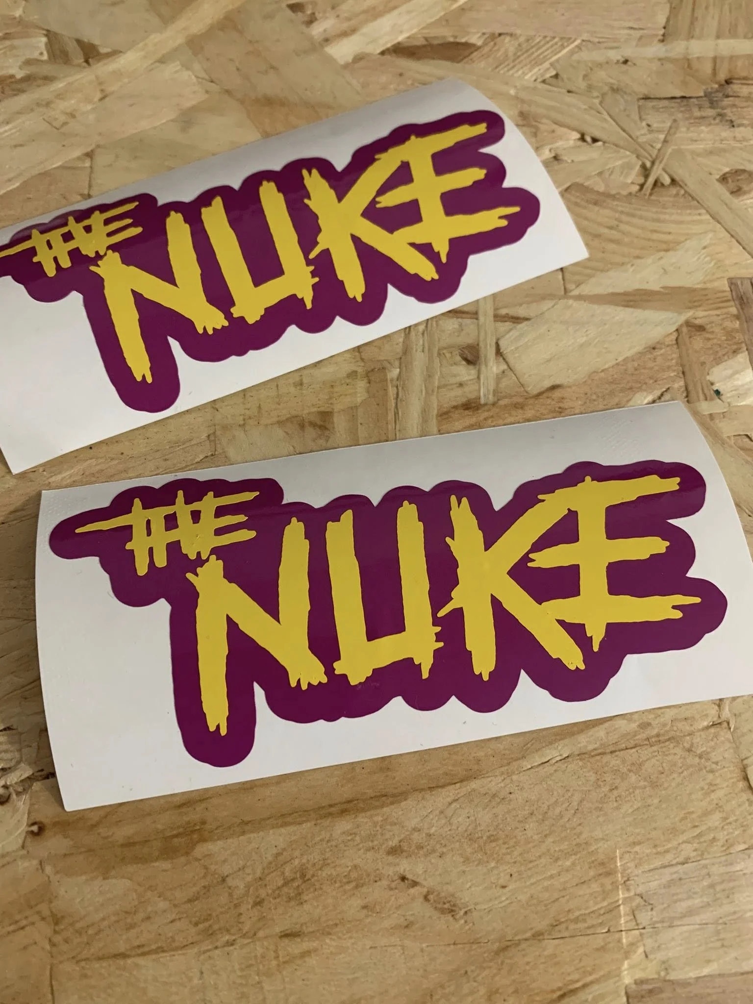 Sticker with the words 'The Nuke' in yellow with purple outline and background on a wood surface.