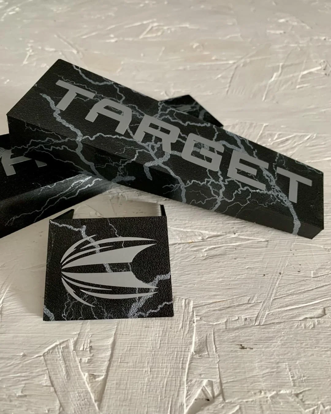 A black box with the word 'TARGET' printed on it and a black sticker with a white emblem, both featuring a lightning bolt design, are placed on a textured white surface.