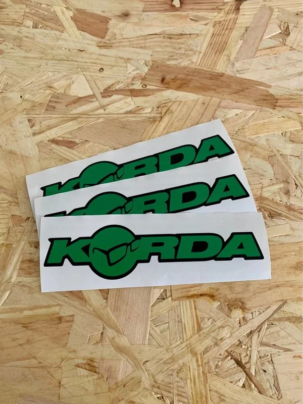 Three KORDA stickers with black text and a green circle with a black outline and a fish silhouette in the center, placed on a wooden surface.