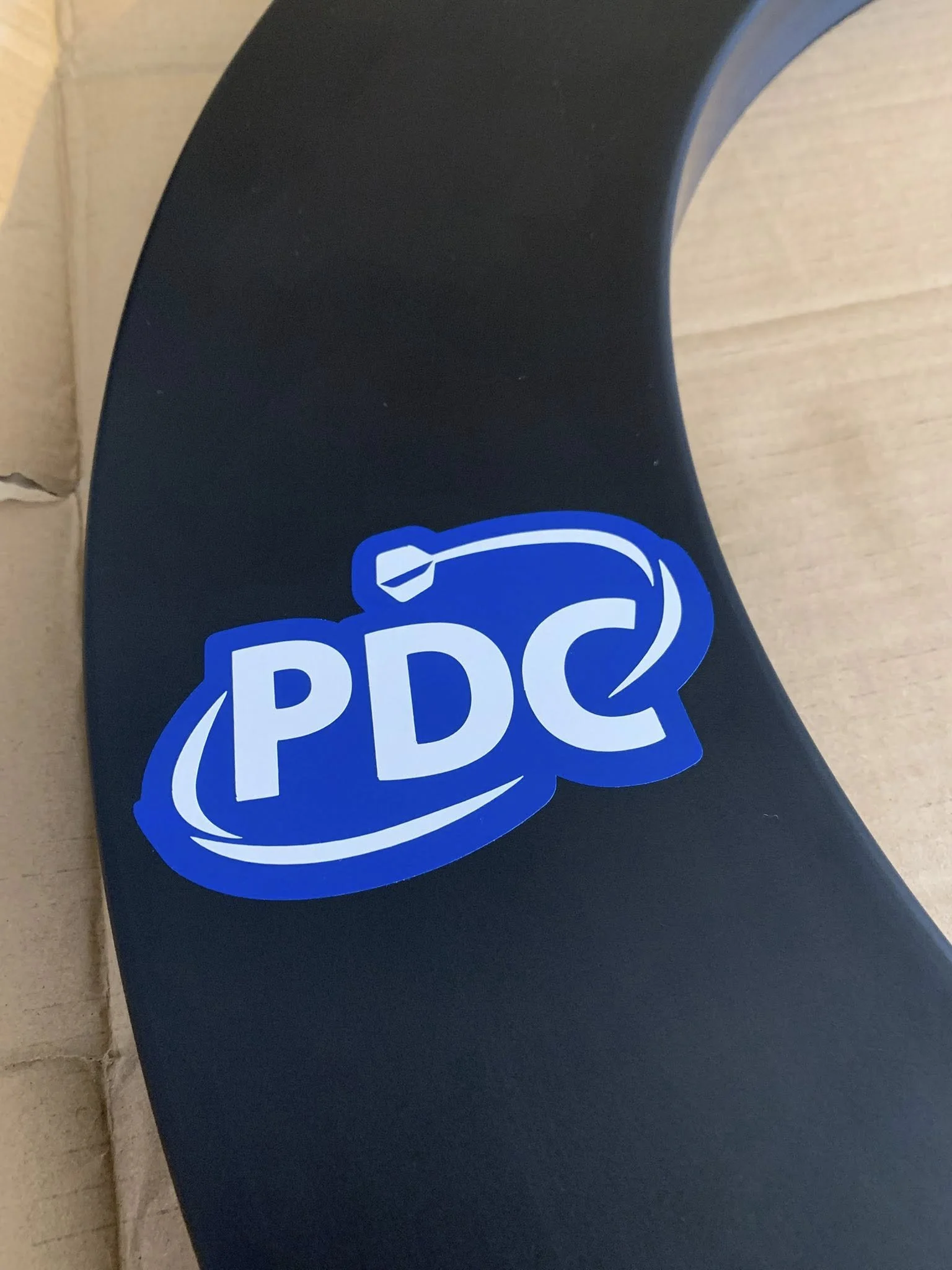 Close-up of a black object with a blue and white PDC sticker, likely part of a vehicle, against a light-colored background.