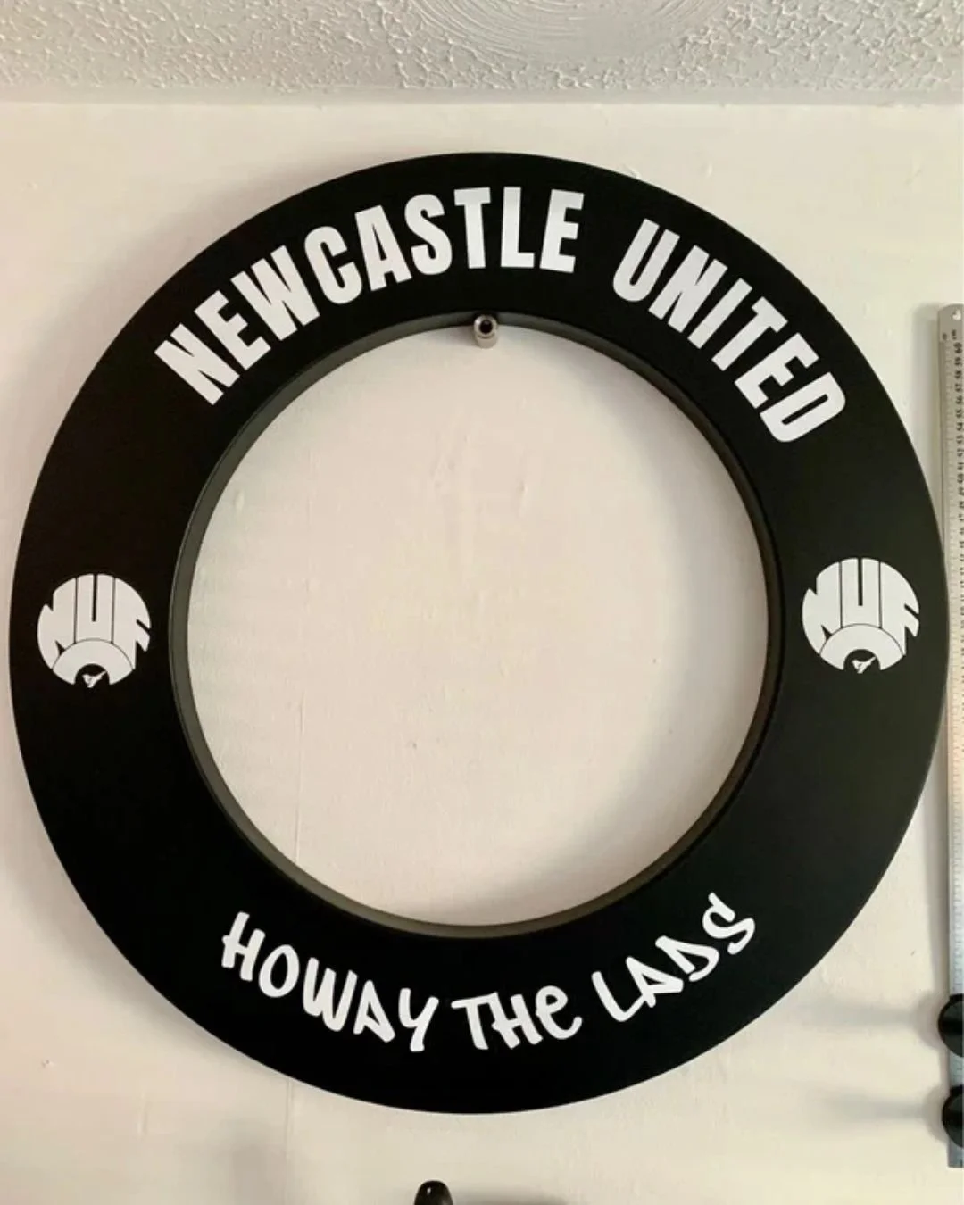 A black circular sign with white text reading "Newcastle United" at the top and "Howay The Lads" at the bottom, with a small graphic of a goalkeeper glove on each side.