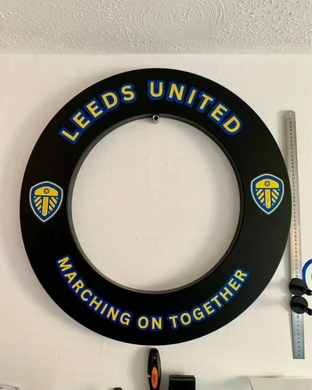 Decorative wall clock with the Leeds United football club logo, the phrase "Leeds United" at the top, and "Marching on together" at the bottom, mounted on a white wall. A measuring tape is on the right side of the clock.