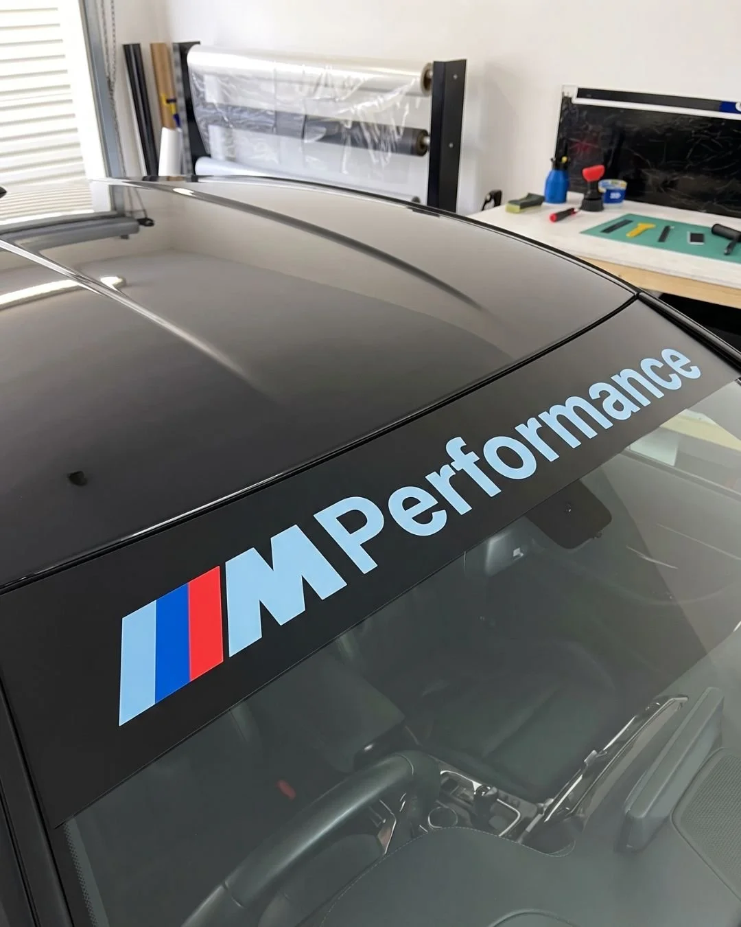 Close-up of a black car windshield with a BMW M Performance decal, in a garage with work tools and equipment in the background.