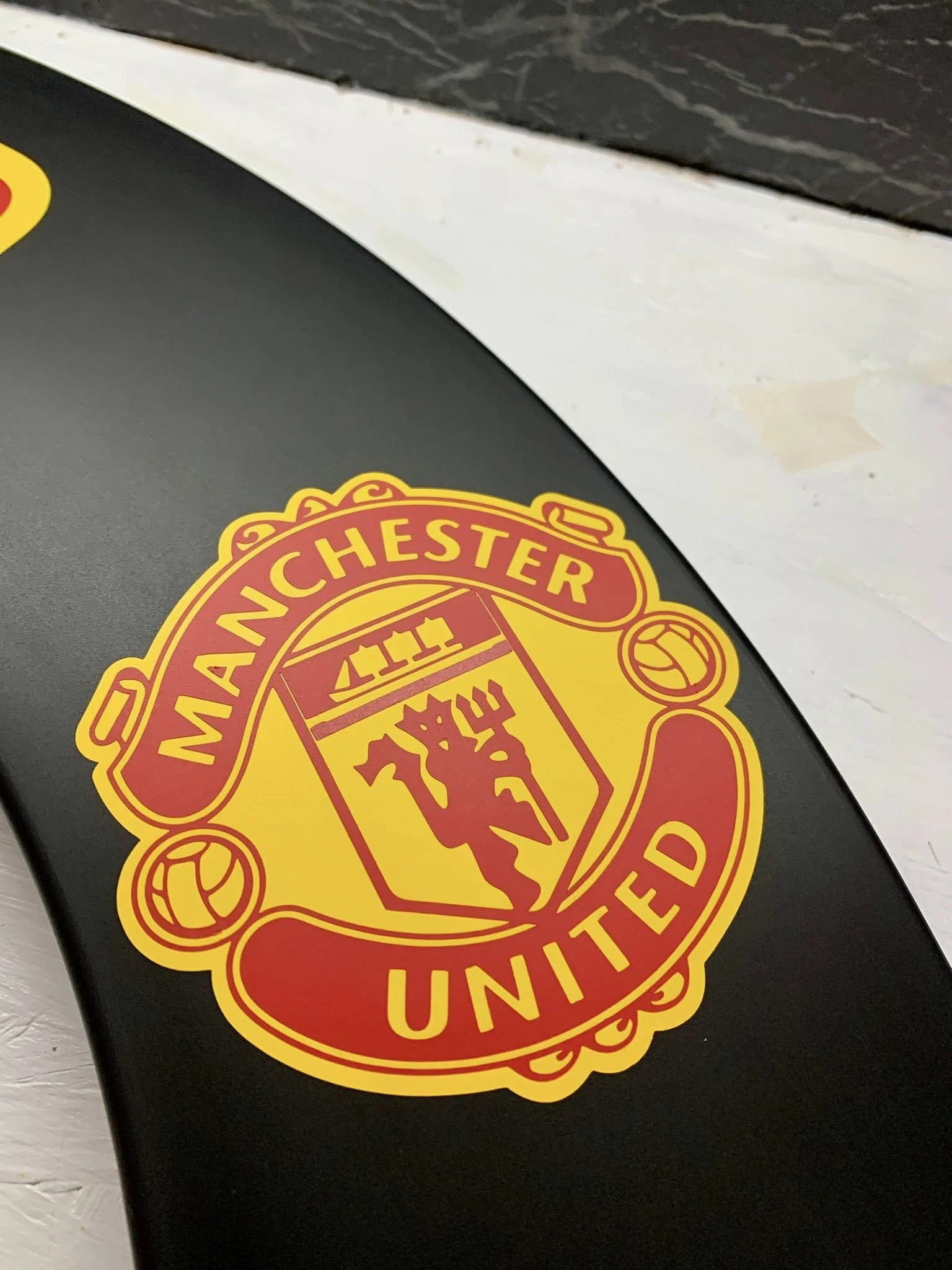 Close-up of a black surface with a Manchester United logo sticker on it. Part of the background shows a black marble surface and a white edge.