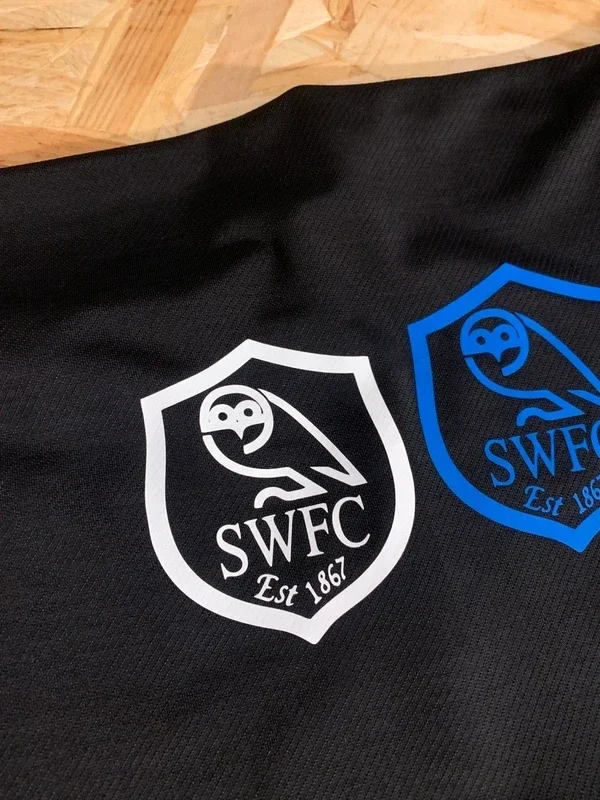 Close-up of a black fabric with two embroidered logos featuring a stylized owl, the letters 'SWFC,' and 'Est 1867.' One logo is white, the other is blue.