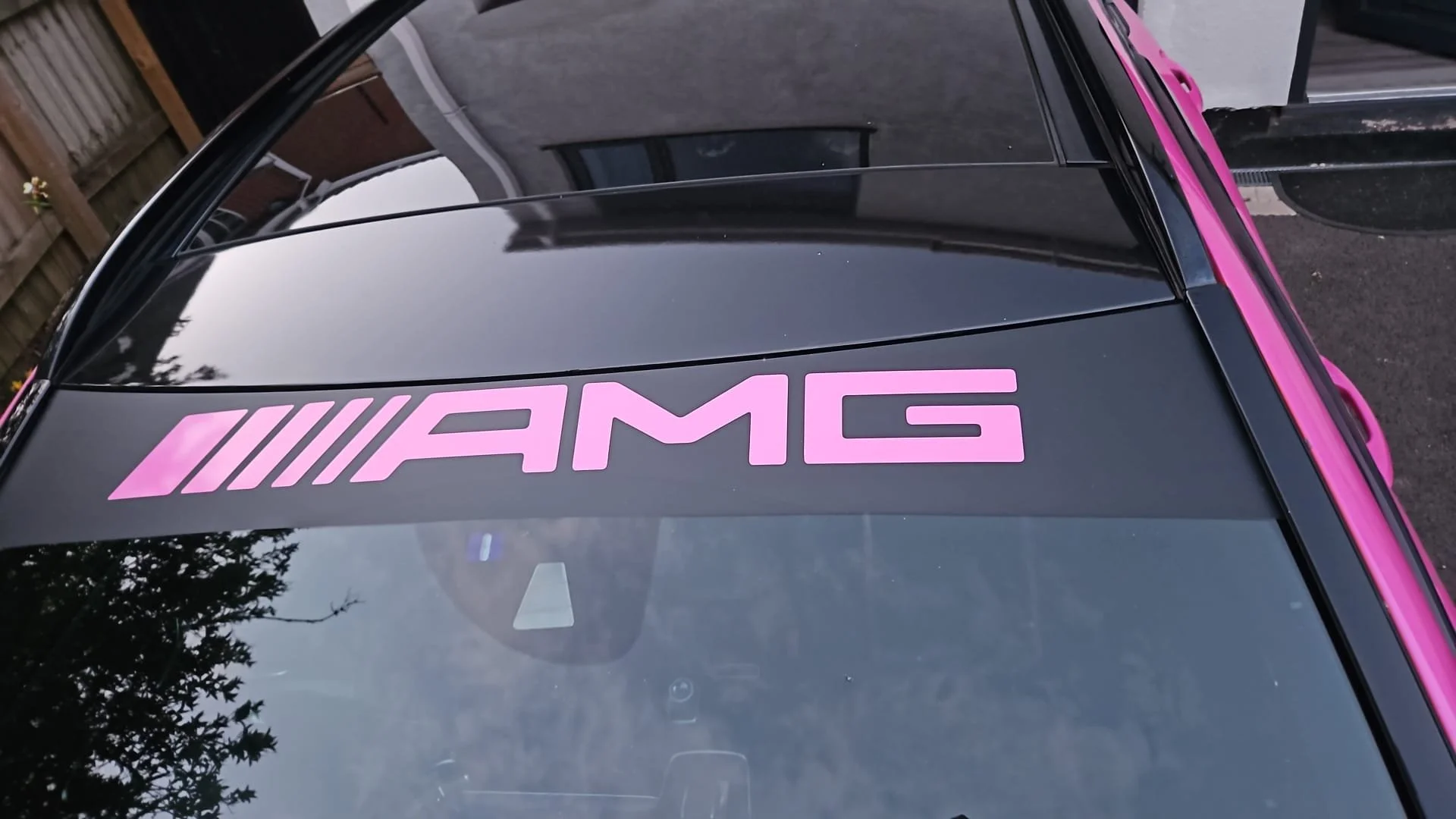 View of a black car with a pink AMG logo on the windshield, parked in a driveway next to a wooden fence and a building with grey walls.