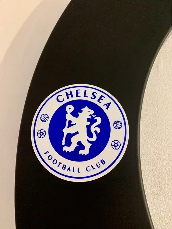 Close-up of a Chelsea Football Club sticker on a black surface, displaying the club's blue and white logo with a lion holding a staff, surrounded by footballs and flowers.