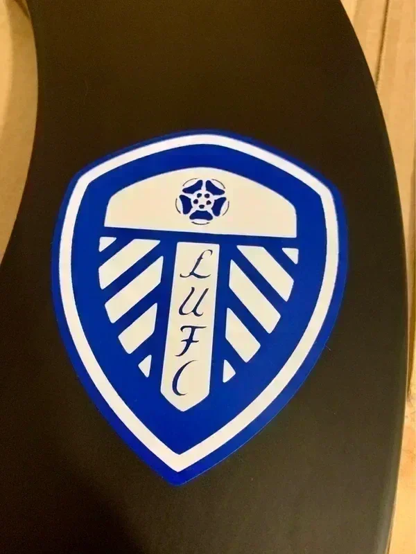 A Leeds United Football Club (LUFC) emblem sticker on a dark surface, featuring a shield with a white and blue design.