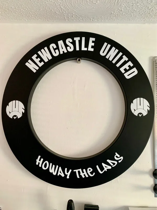 A black circular wall sign with white text reading "Newcastle United" at the top and "Howay the Lads" at the bottom. The sign has two logos on either side featuring stylized fists.