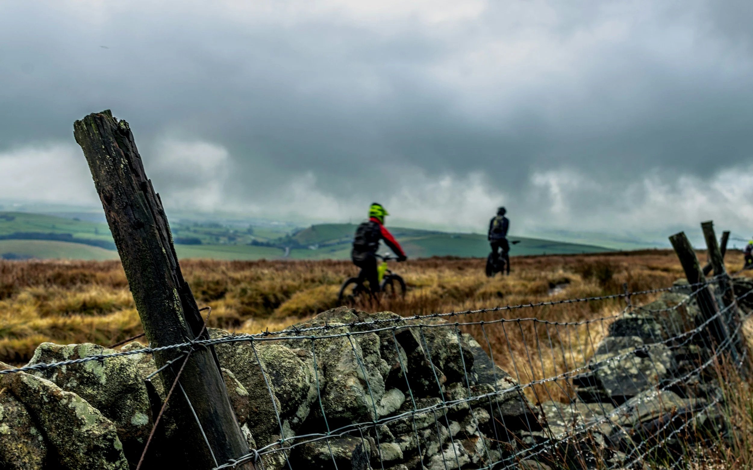 Two children riding bikes along a rural trail, with an old wooden fence and stone wall in the foreground, and a cloudy sky over rolling green hills in the background.