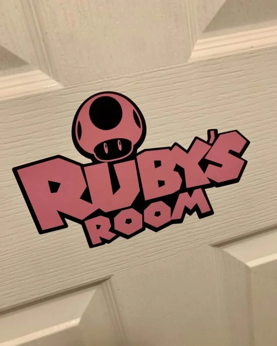 A sticker on a door with the words 'RUBIK'S ROOM' in bold, pink, and black text, featuring a cartoonish pink mushroom with black spots above the text.