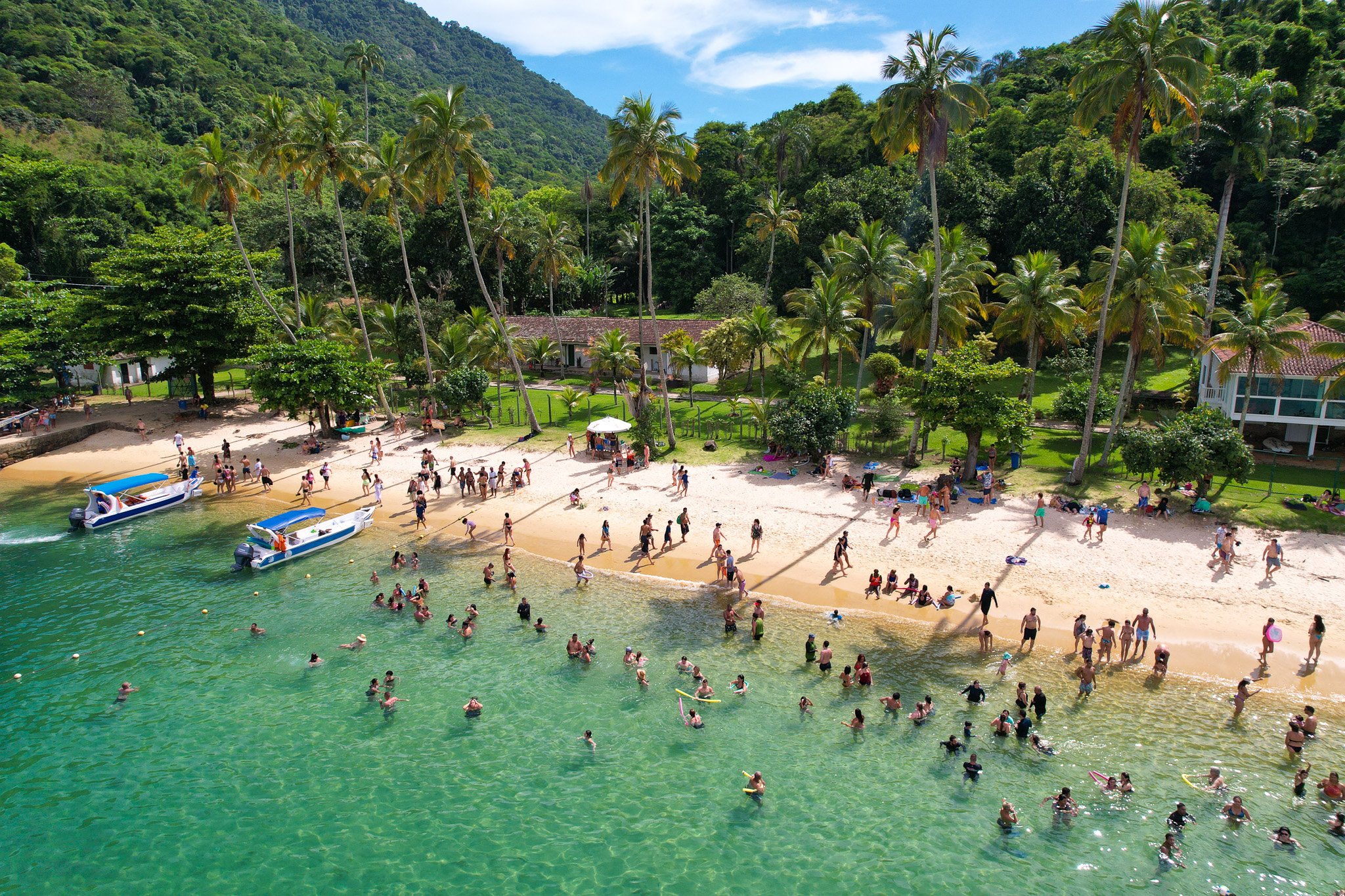Aerial view of a beach with many people swimming in turquoise water and walking on the sandy shore, surrounded by lush palm trees and greenery. Two small boats are anchored near the shore.