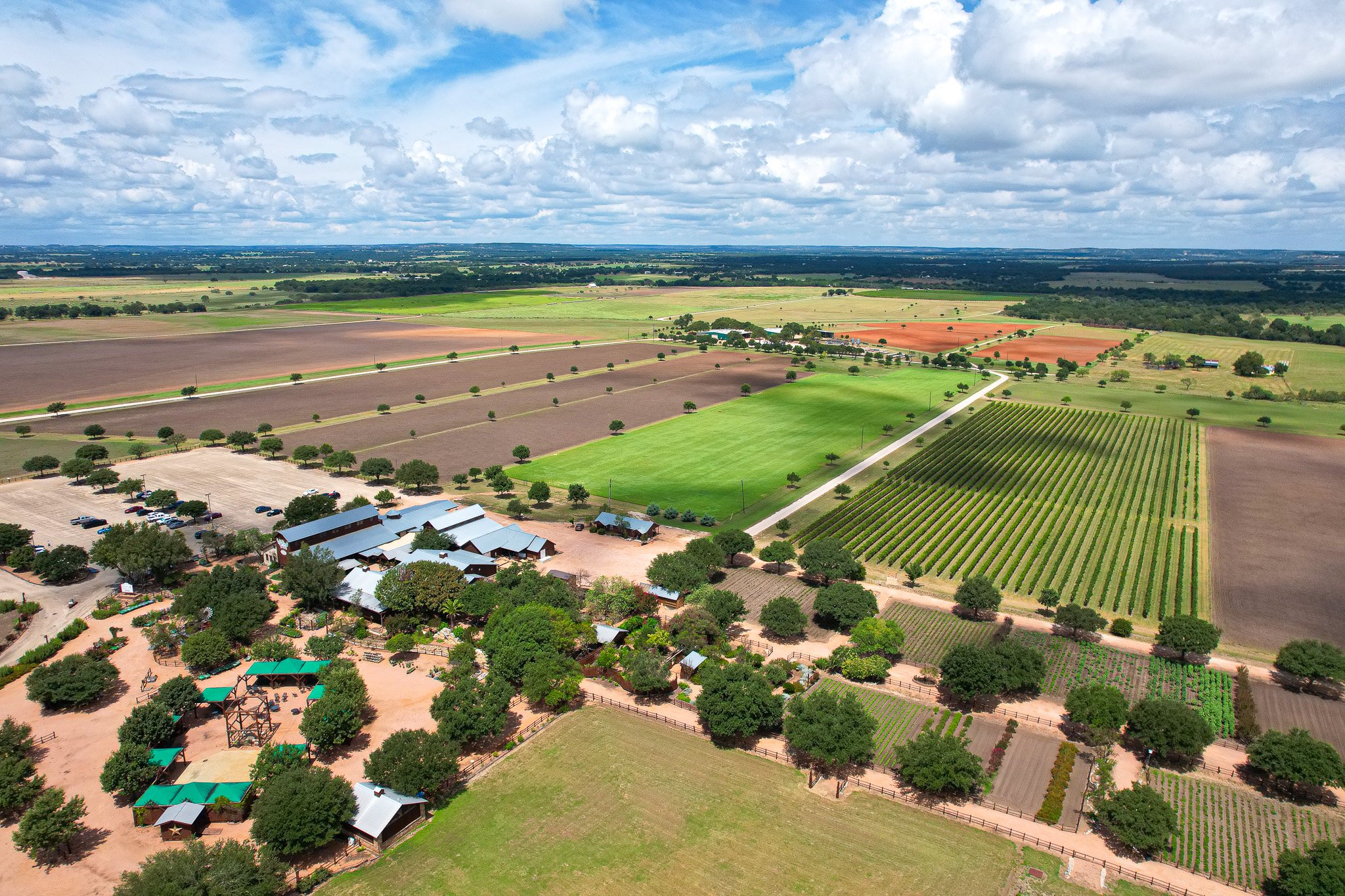 Aerial view of a large vineyard and farmlands with rows of cultivated crops, surrounded by green fields and trees. Several buildings are visible, along with a parking area and a road running through the landscape. The sky is partly cloudy.