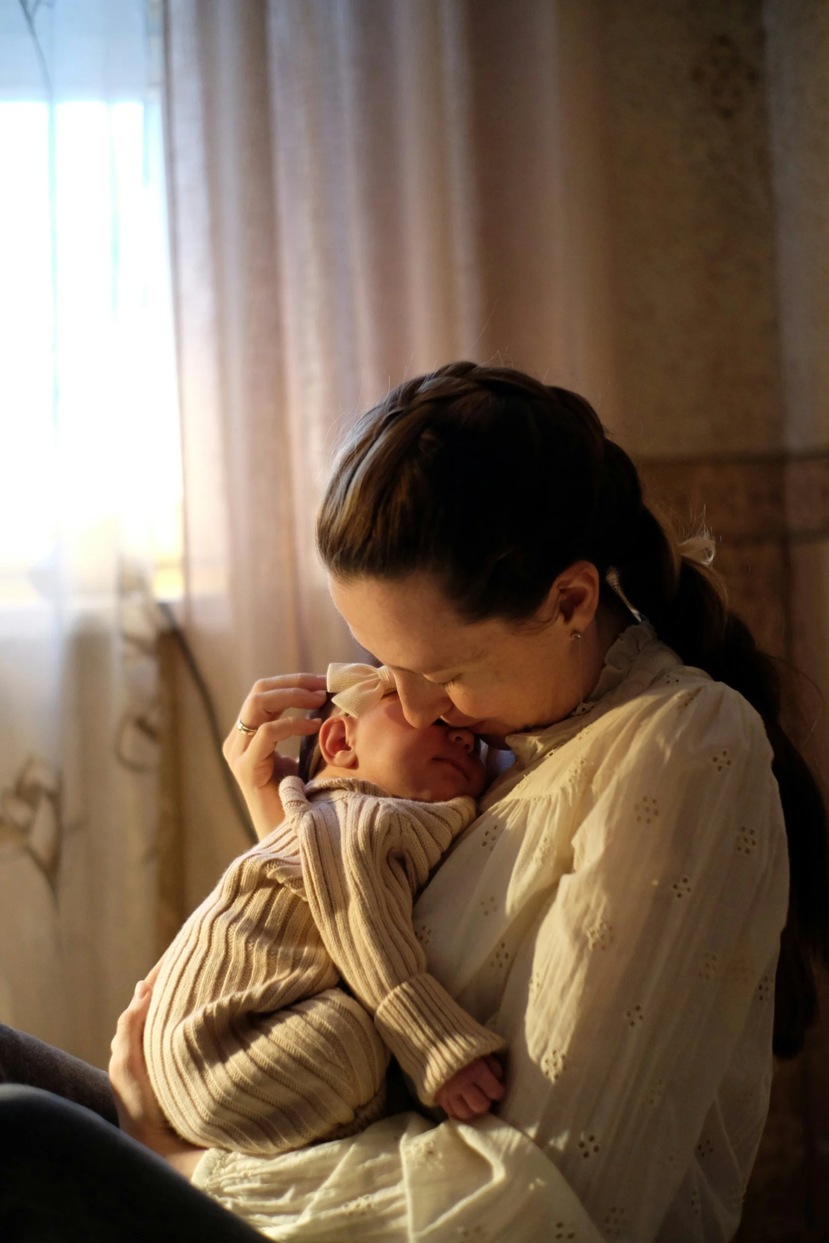 A woman with brown hair, wearing a cream-colored dress, holding a young girl with a bow in her hair, sharing a tender moment with the child resting her head on the woman's chest in a warmly lit room with curtains in the background.