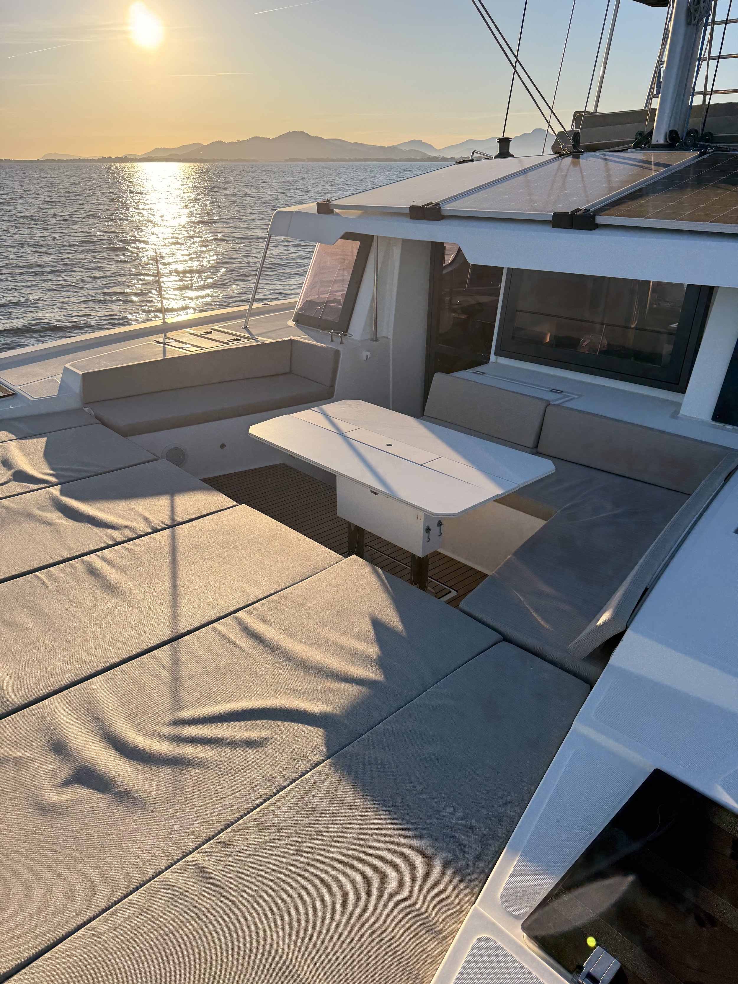 Une partie du pont d'un catamaran Bali 4.6 avec des coussins, une table pliante, une vue sur la mer et la lumière du coucher de soleil.