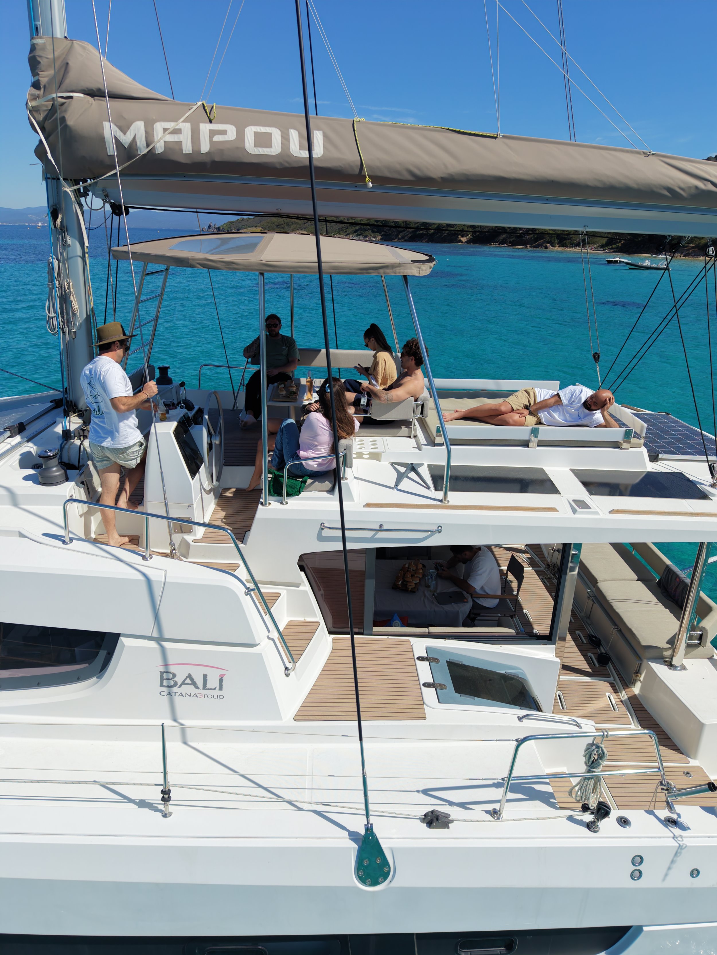 Un groupe de personnes sur un catamaran Bali 4.6 en Méditerranée, avec des eaux turquoises, un ciel bleu clair, et une côte rocheuse en arrière-plan. Certains personnes sont assises autour d'une table, d'autres sont allongées sur le pont.