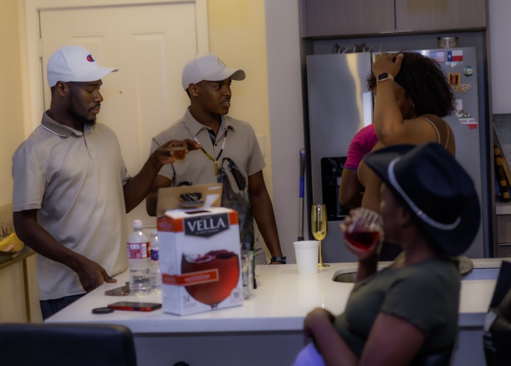 Three men and two women in a kitchen. Two men are standing by the counter, one holding a glass of drink. The woman in the foreground is sitting and drinking from a glass, wearing a hat. The other woman is standing with her hand on her head, beside a 