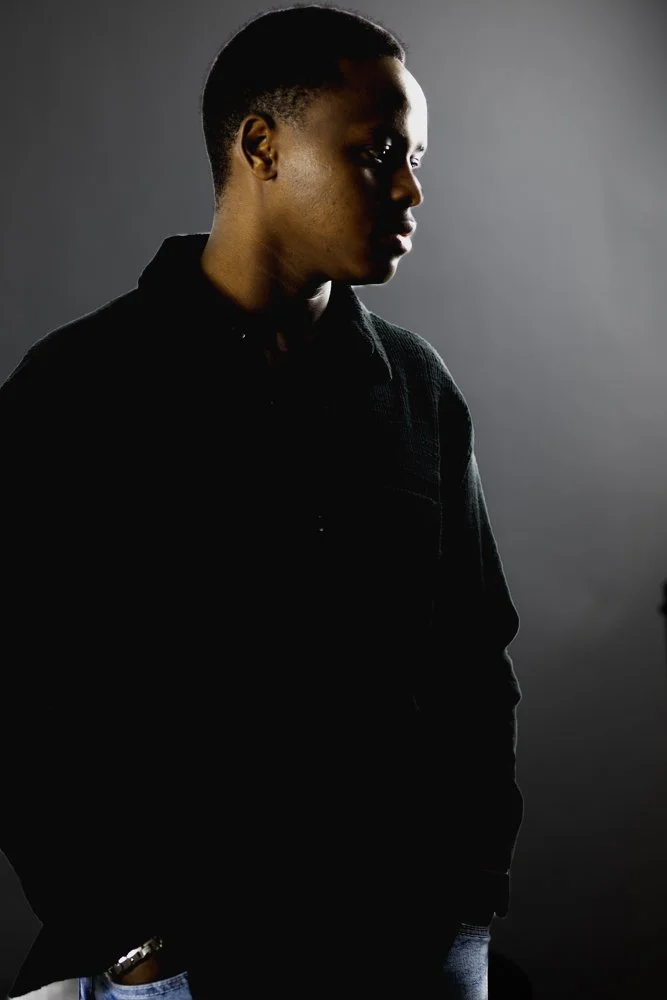 Profile of a young man with dark skin, short hair, and wearing a black jacket, standing against a gray background.
