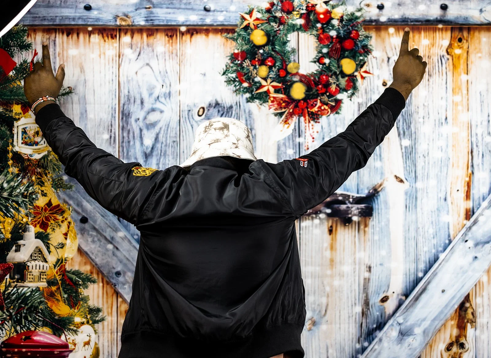 Person with their back to the camera, raising both arms, standing in front of a decorated Christmas wreath on a rustic wood wall.