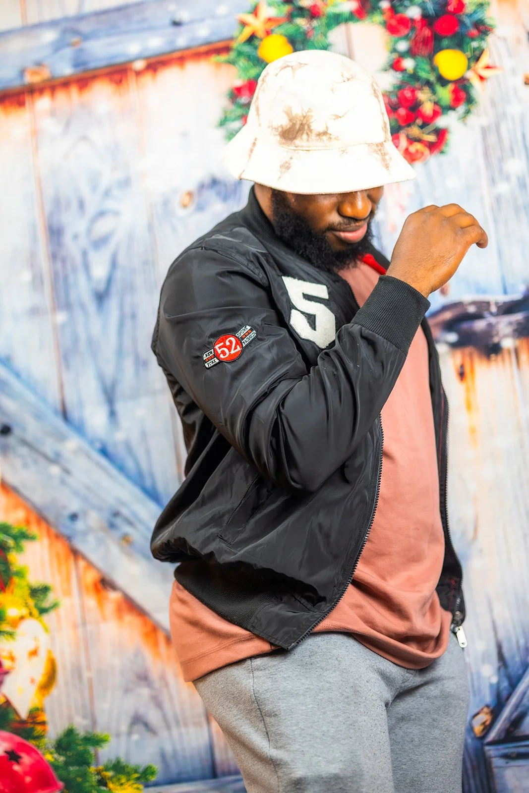 A man in a white bucket hat, black jacket with patches, salmon t-shirt, and gray sweatpants standing in front of a Christmas decorated background.