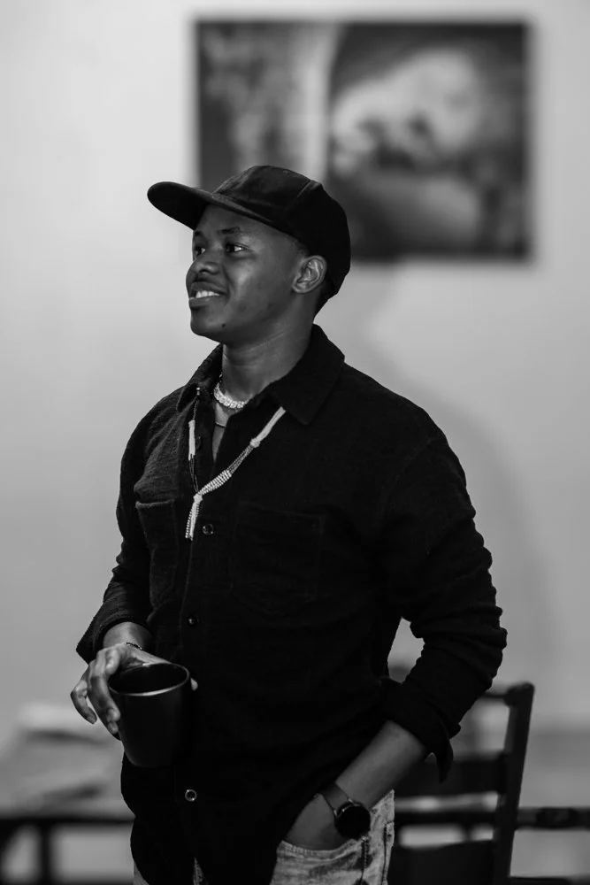 A young man wearing a cap, necklace, and dark jacket, holding a mug while smiling and standing indoors. Blurred artwork is visible in the background.
