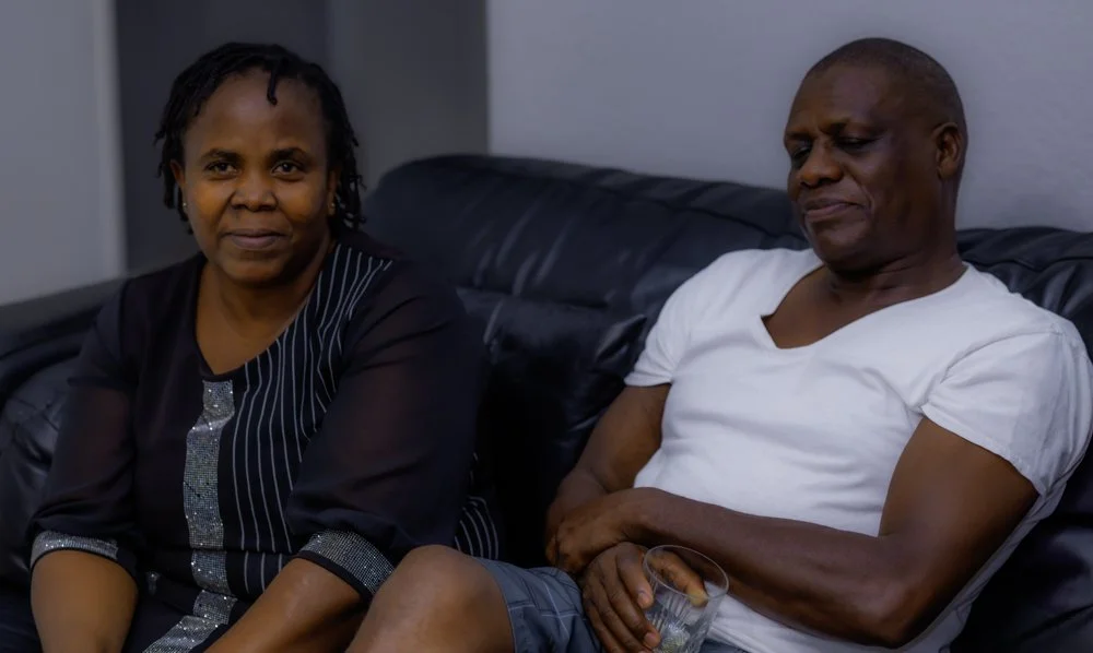 A middle-aged woman and an older man sitting on a black leather couch. The woman has short dreadlocks and is wearing a black top with silver details, looking at the camera. The man is wearing a white T-shirt, has his arms crossed, and appears to be l