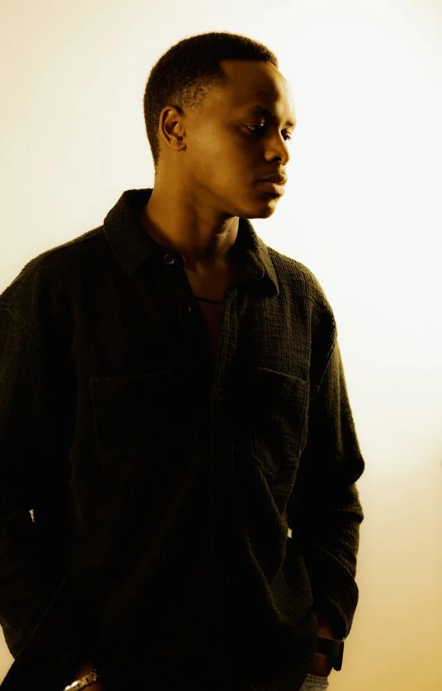 A young man with short hair, wearing a black corduroy shirt, looking to the side with a neutral expression, standing against a plain background.