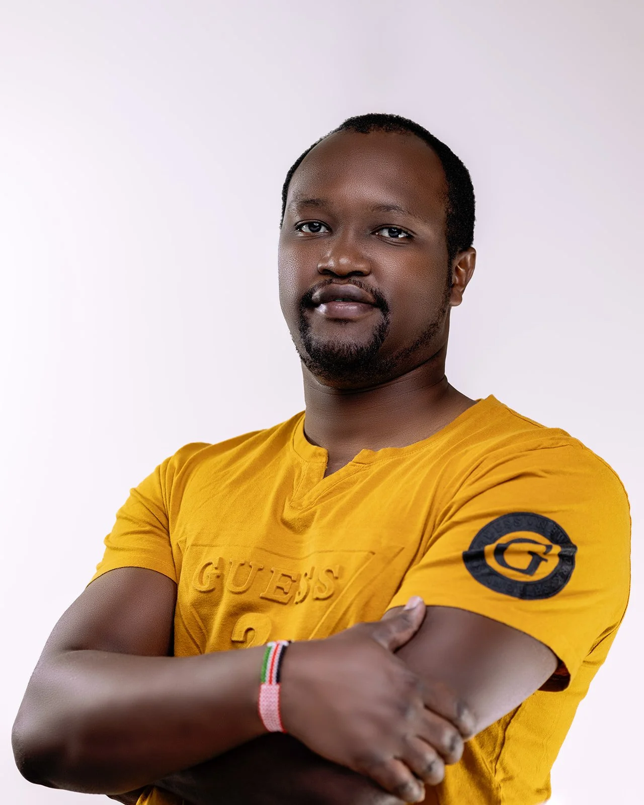 A man with short black hair, a goatee, and a mustache wearing a yellow Guess T-shirt with a black G logo on the left sleeve, crossing his arms, and standing against a white background.