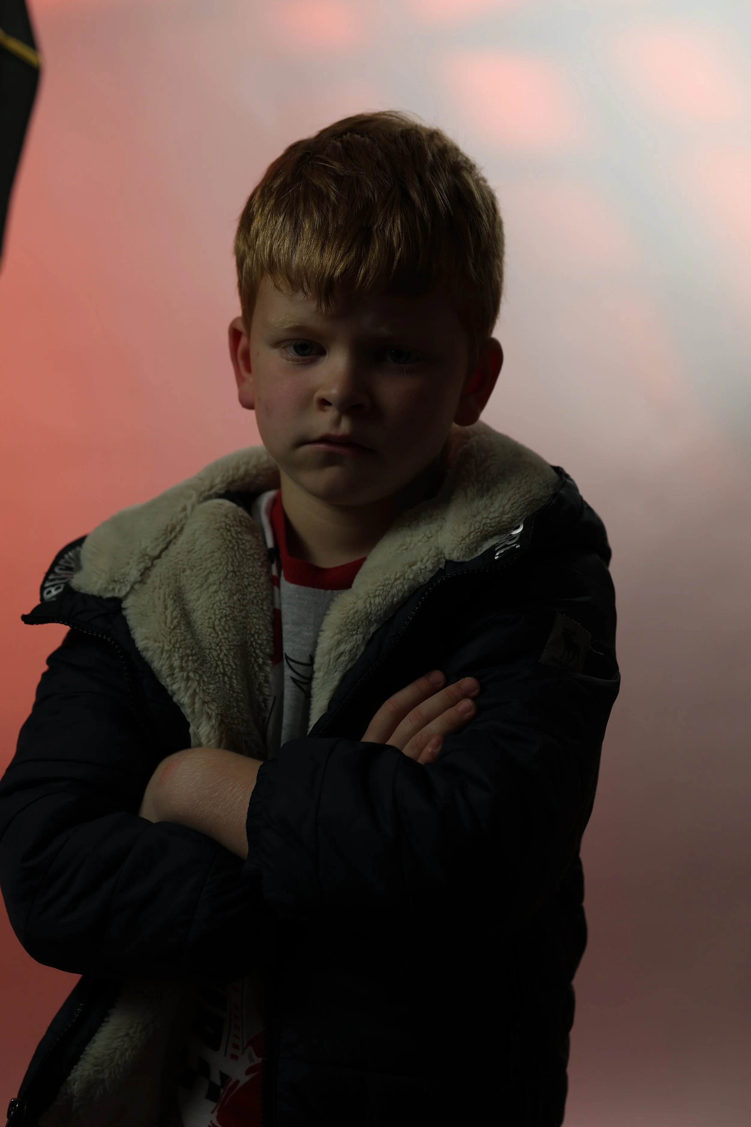 A young boy with crossed arms, looking serious or upset, standing in front of a pinkish background.