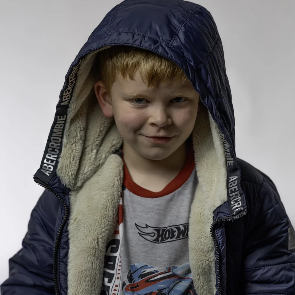 A young boy with red hair and blue eyes wearing a dark blue hooded jacket with a beige fleece lining, and a gray T-shirt with colorful graphics, looks at the camera against a neutral background.