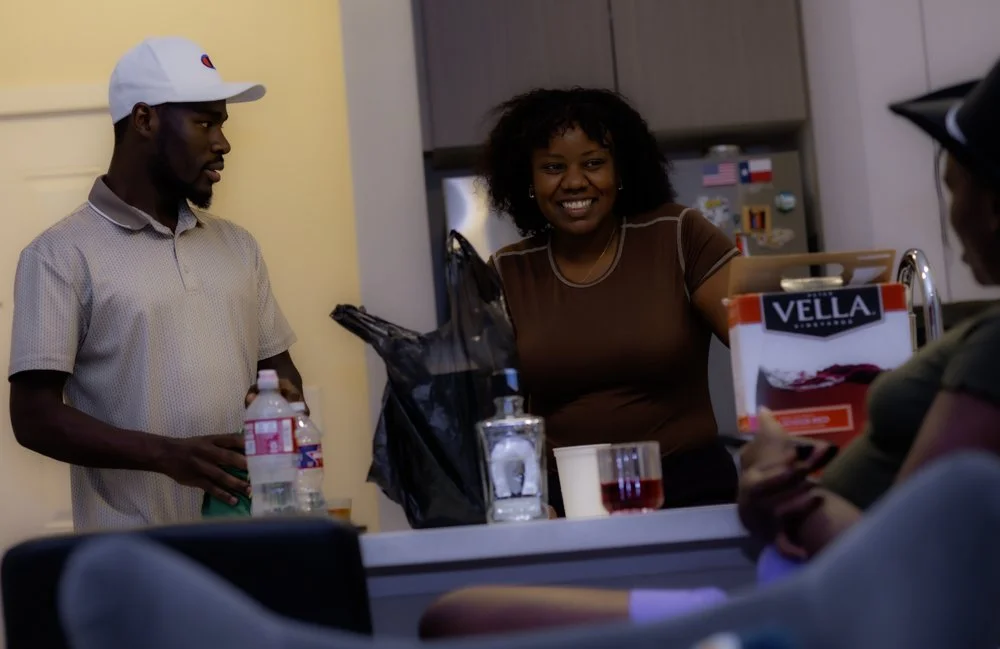 Three people gather around a kitchen island, one woman smiling and talking, one man holding a water bottle, and another woman with sunglasses, partially visible.