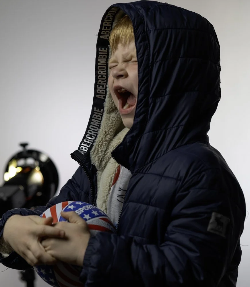 A young boy with blond hair, wearing a navy blue puffer jacket with the hood over his head, is shouting or yawning with his eyes closed, holding a basketball decorated with an American flag design.