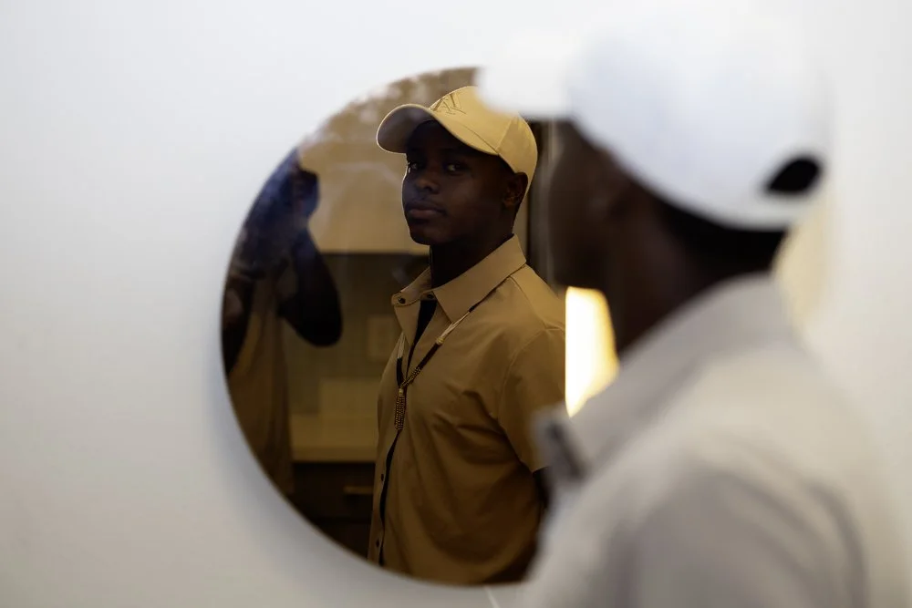 A young man in a tan baseball cap and shirt is seen through a round mirror, looking at and focusing on another person.