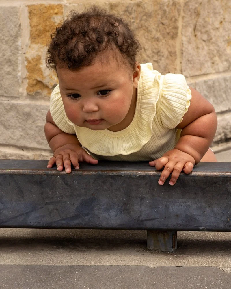 A young child with curly hair in a yellow outfit crawling on a black surface against a brick and stone wall background.