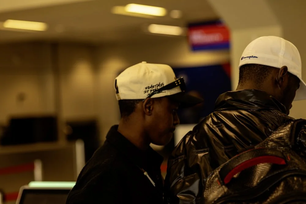 Two men at an airport check-in counter, both wearing white baseball caps and dark jackets.
