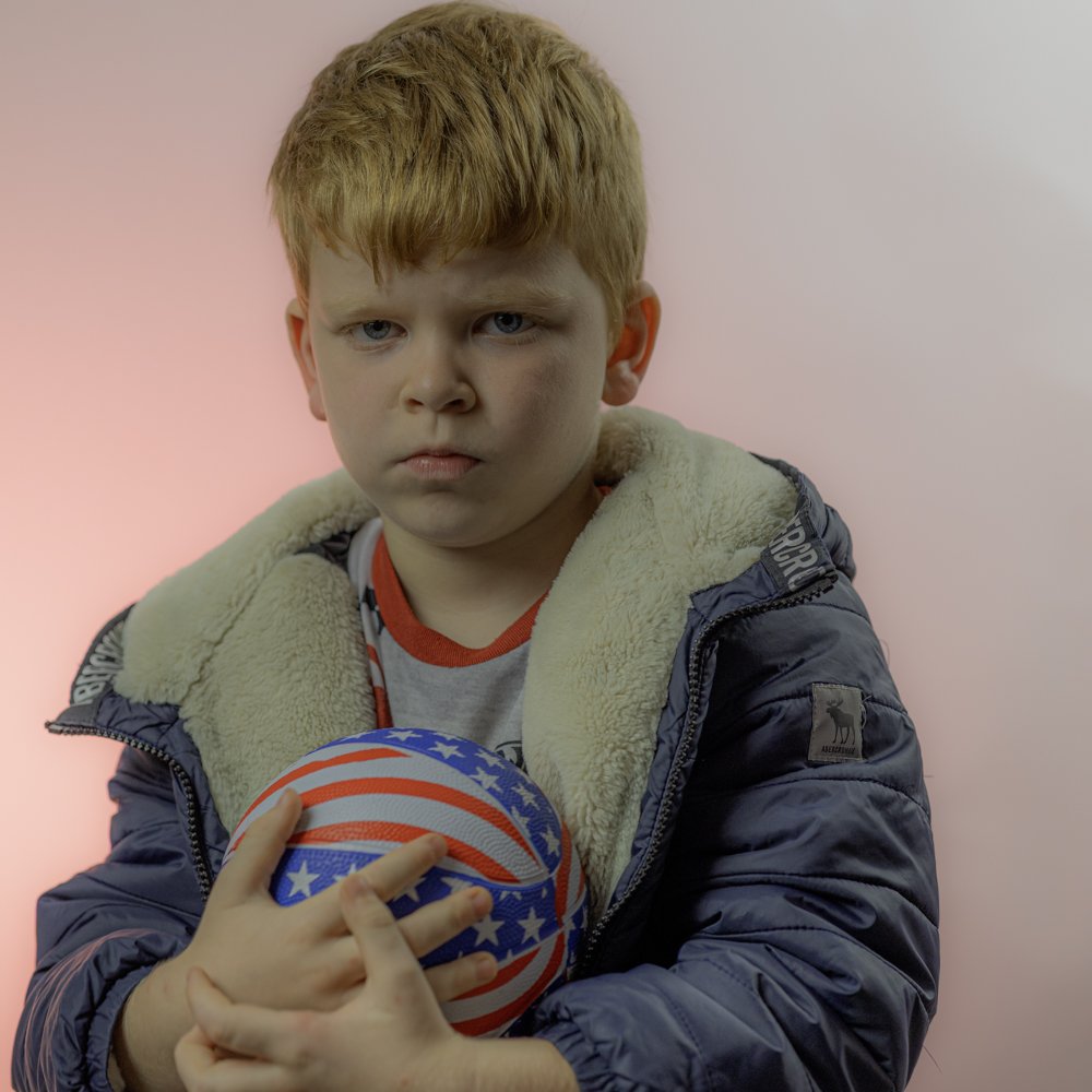 A young boy with red hair, wearing a jacket with beige lining, holding a ball with red, white, and blue star stripes, standing against a pink background.