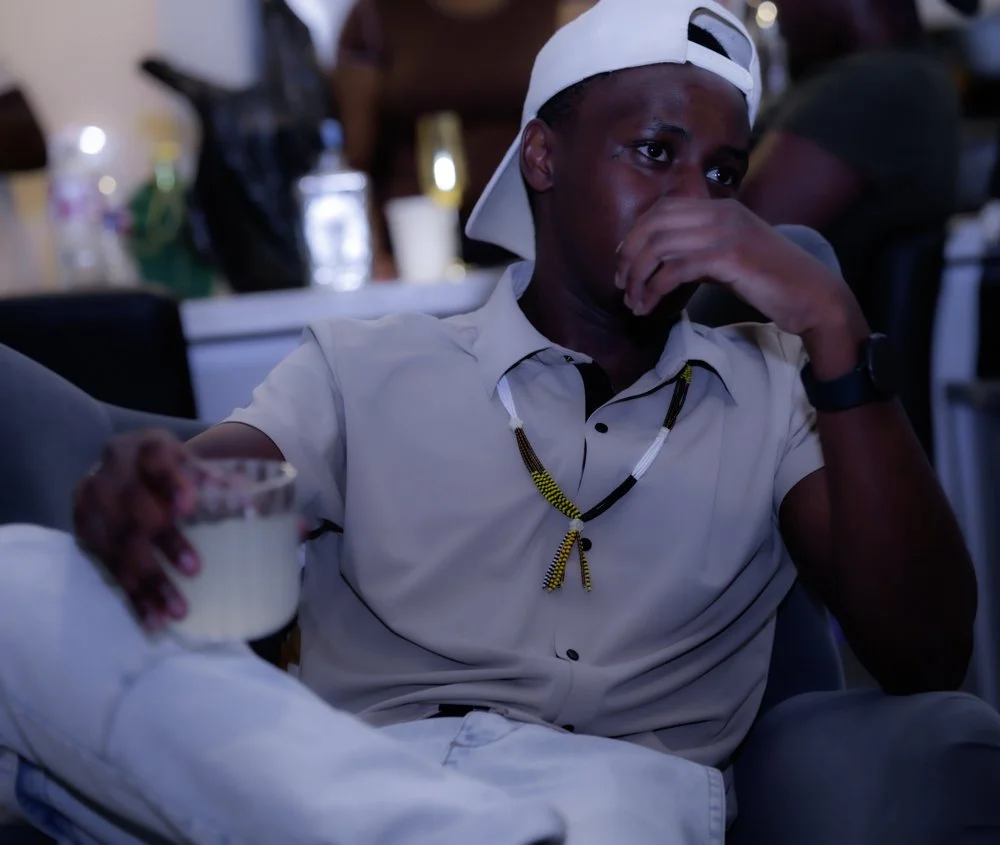 A young man with a dark complexion wearing a white shirt and a backward white cap, sitting at a table holding a bowl of food, with a thoughtful or emotional expression on his face.