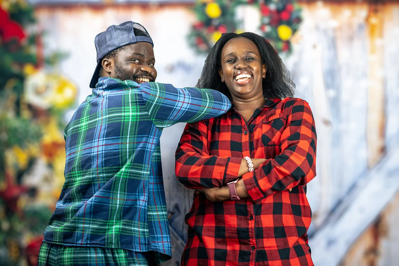 A smiling man in a blue plaid shirt and baseball cap playfully pokes a woman in a red and black plaid shirt, who is laughing with her arms crossed.