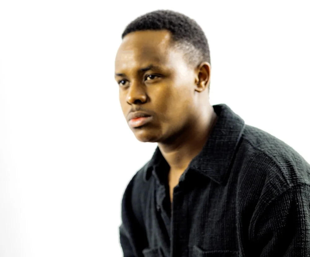 A young man with dark skin and short hair wearing a black textured shirt, looking slightly to the side against a plain white background.