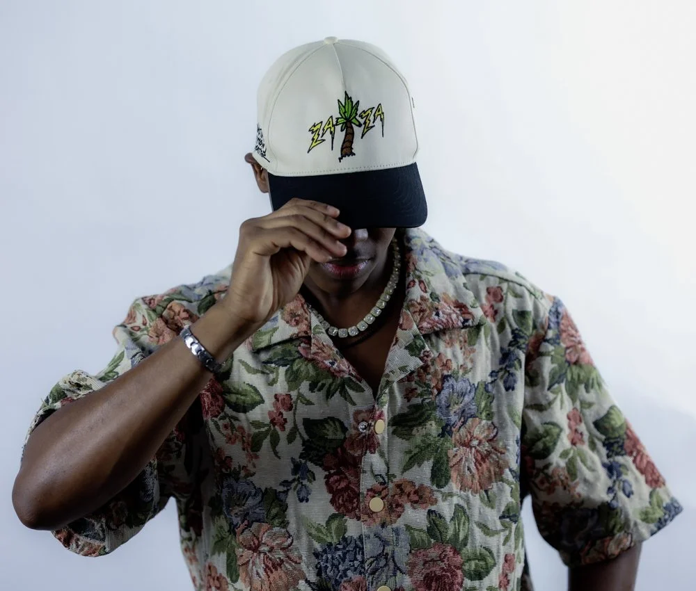 Person wearing a floral shirt and holding a baseball cap with a marijuana leaf design and lightning bolts on it, partially covering their face.