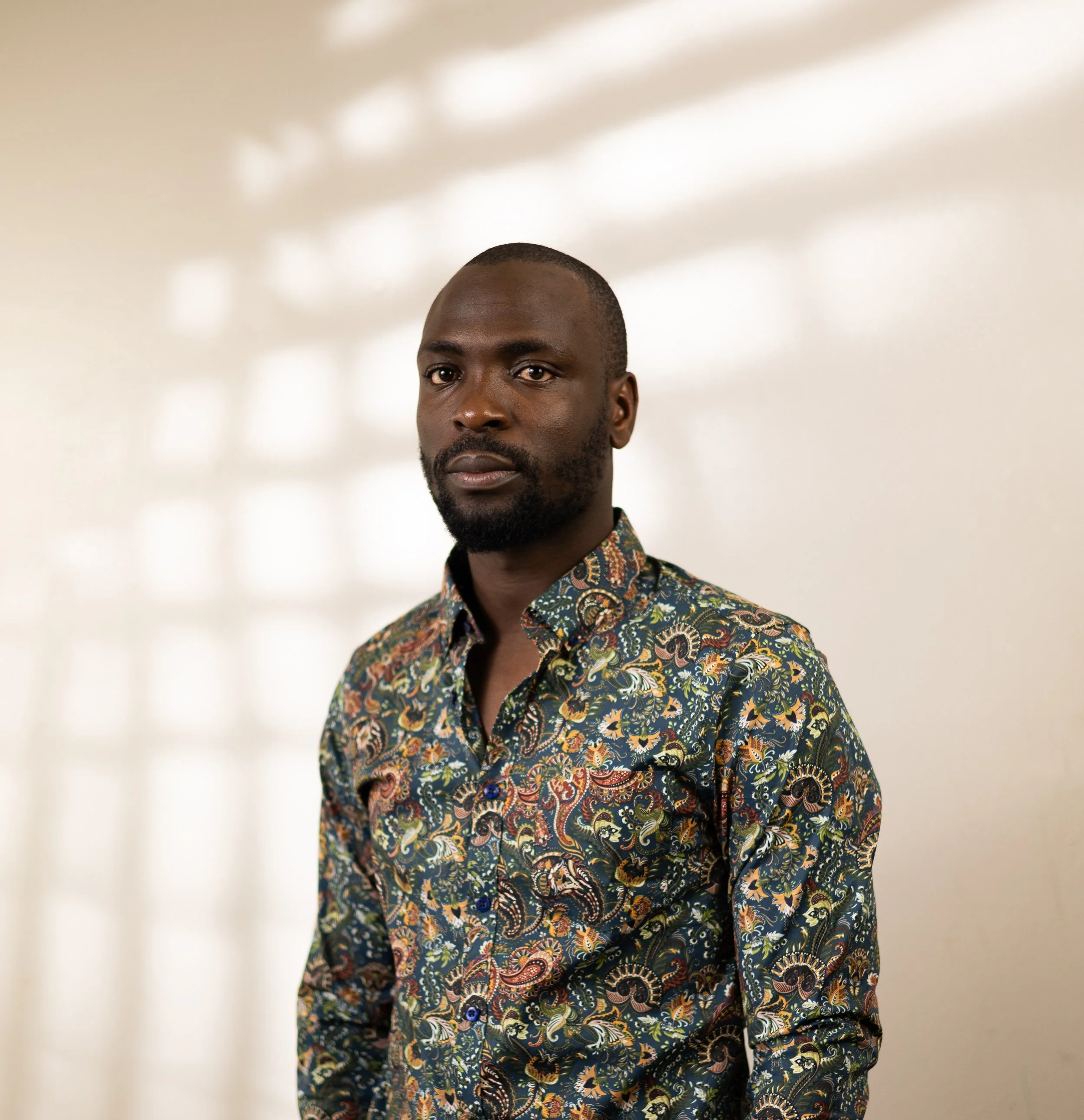 A man with dark skin and short hair wearing a colorful, patterned button-up shirt standing indoors with a plain white wall and soft light shadows in the background.
