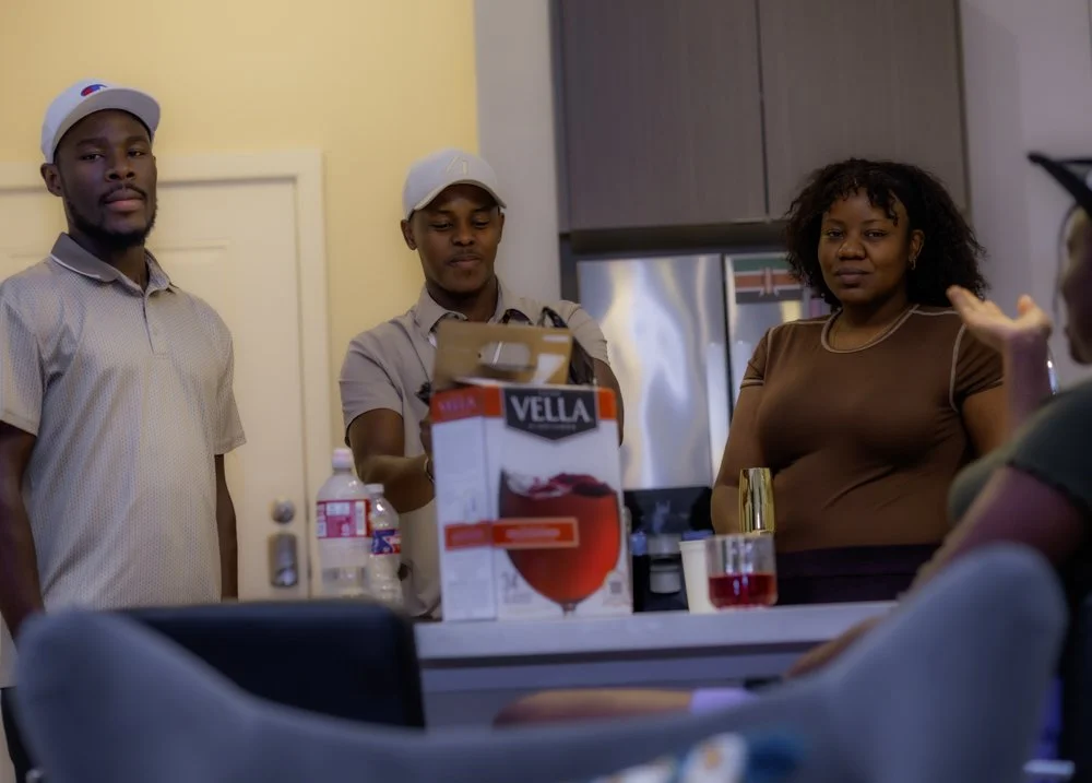 Three people standing in a kitchen, two men and a woman, with a third person seated at the counter. The man on the left wears a light-colored polo shirt and a white cap, the man in the middle wears a beige uniform and a white hat, and the woman on th