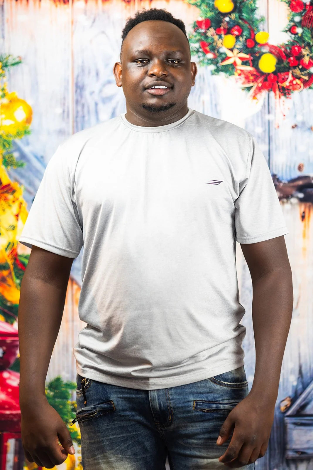 A man standing in front of a Christmas-themed background with wreaths and ornaments, wearing a light gray t-shirt and jeans.