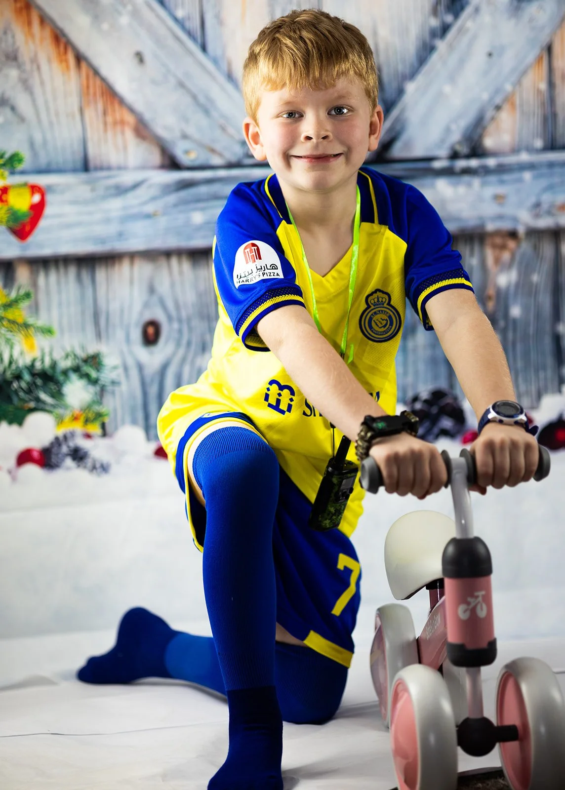 A young boy with red hair riding a pink and white bicycle indoors, dressed in a yellow and blue soccer uniform, smiling at the camera.